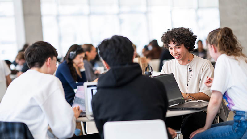 Young adults sit around a table with laptops in a bright study area, smiling and focused.