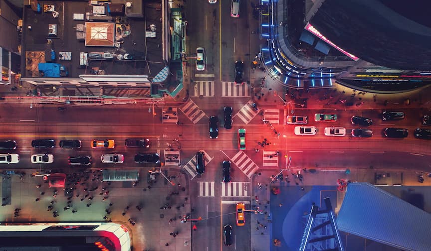 Aerial view of a busy city intersection at night with red and blue lights, cars, and pedestrians moving.