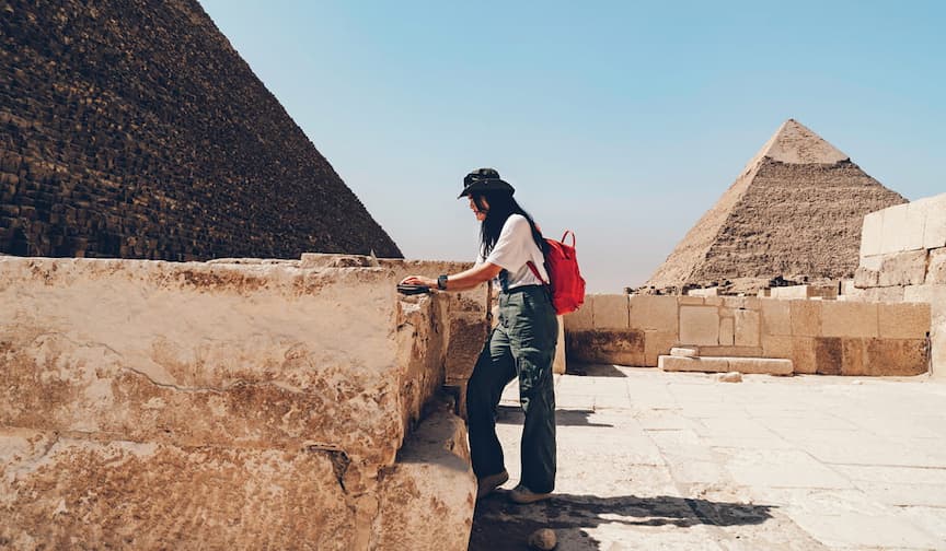 A person with a red backpack explores the Pyramids of Giza under a clear blue sky.