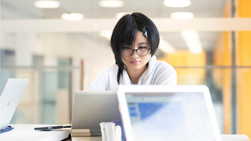 Young person with glasses works intently on a laptop in a bright, modern office.