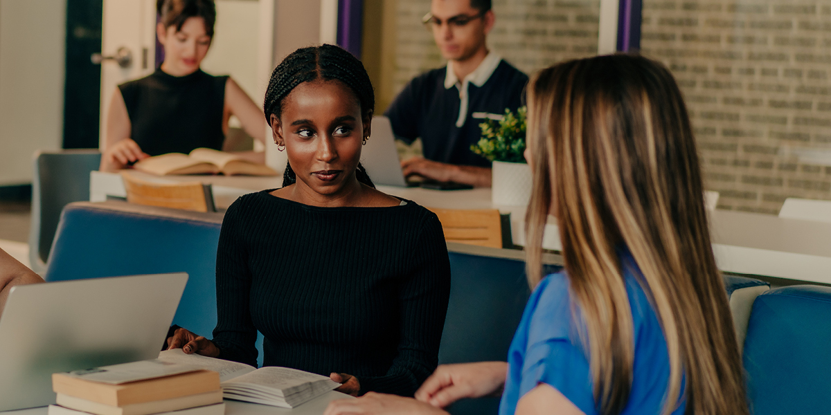 Two women sit in a modern room, focused in conversation; one with braids in black, the other in blue.
