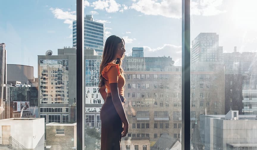 A woman in an orange top stands by large windows, side profile, overlooking a cityscape under clear sky.