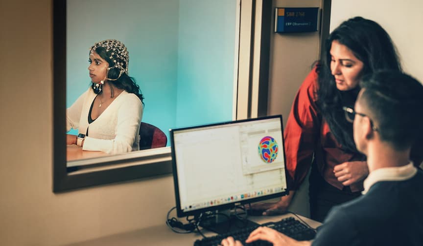 A woman wearing a brain-monitoring cap sits, watching a researcher view data with another person through a window.
