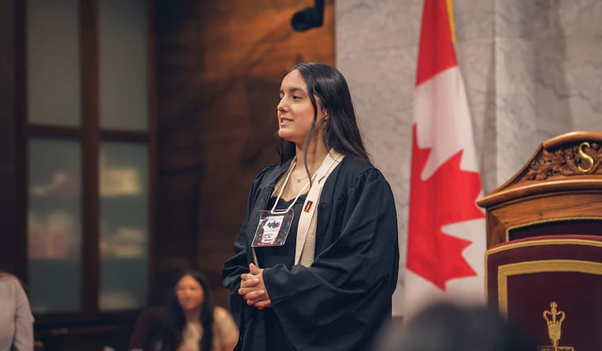 Young woman in a black robe stands confidently, speaking with a Canadian flag in the background.