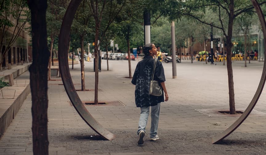 A person walks through a large metal ring on a tree-lined city street, carrying a bag and dressed casually.