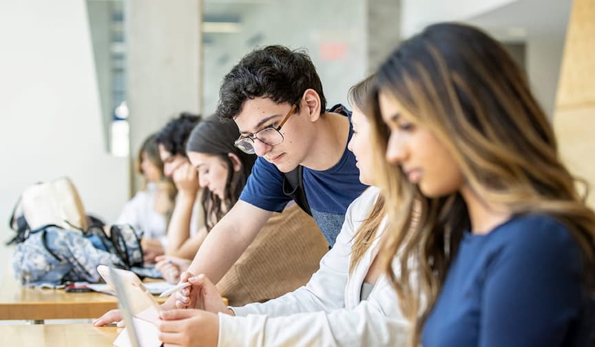Students study at a wooden table in a bright library, using tablets and laptops, focused and engaged.