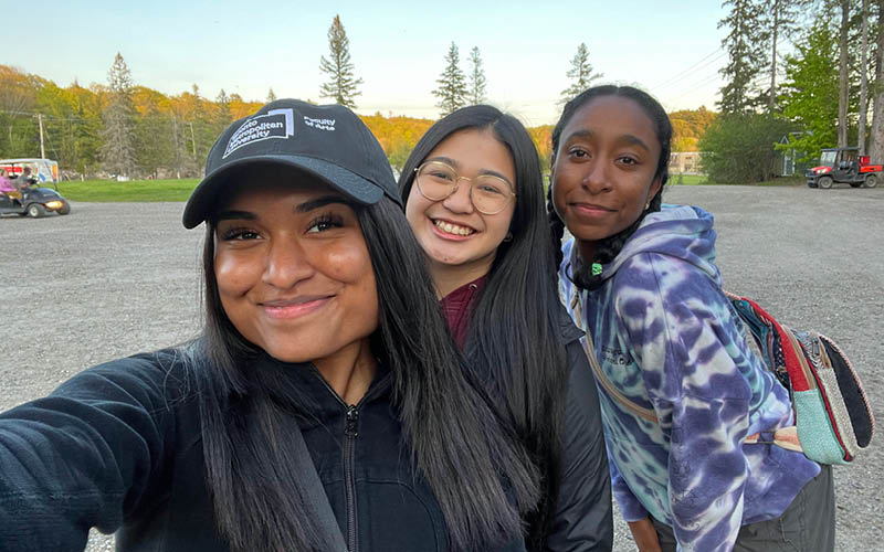 Three friends smile outdoors in gravel, surrounded by trees. One wears a black cap, another glasses, and the third a tie-dye hoodie.