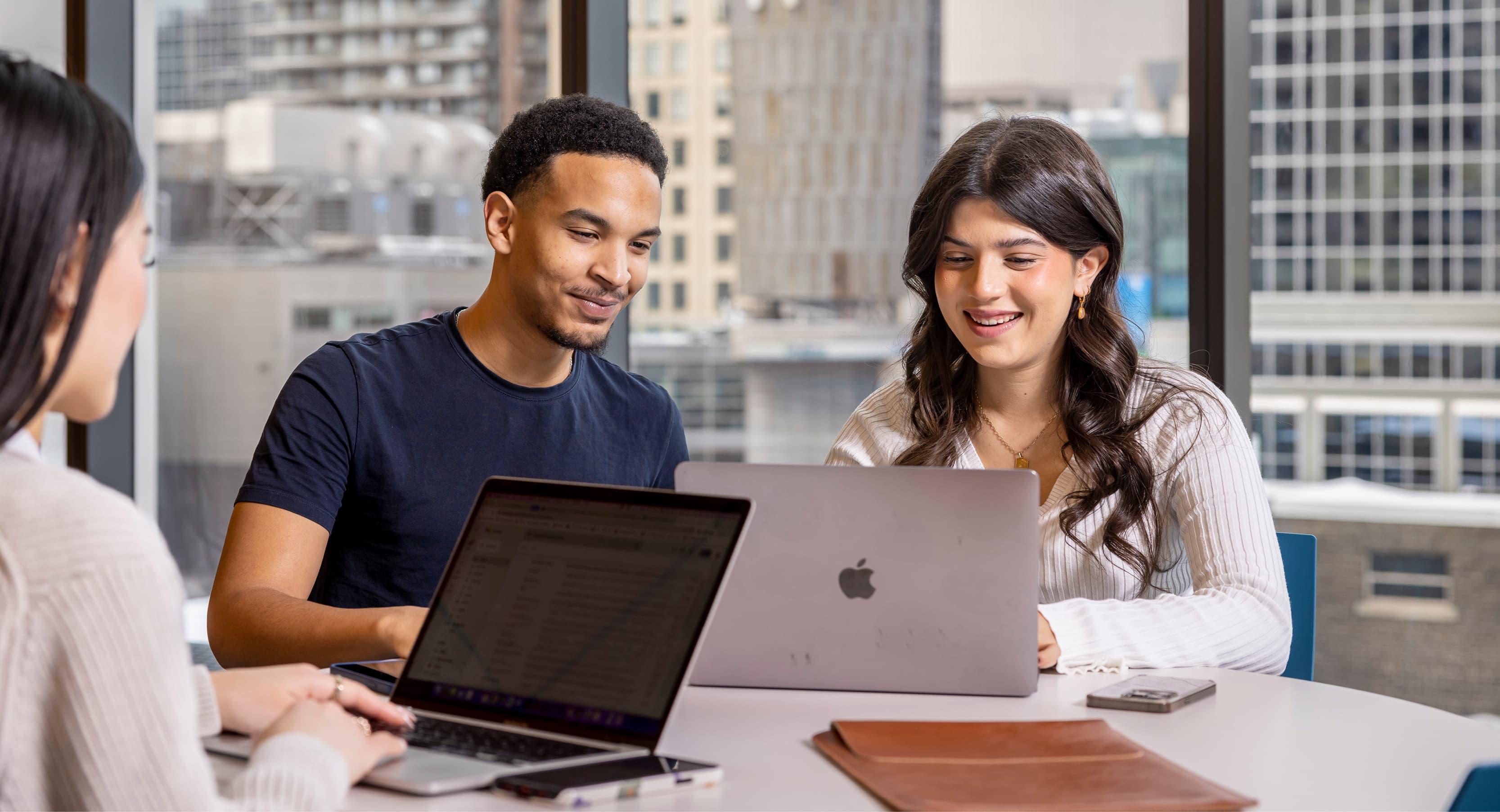 Two individuals sit at a table with laptops, smiling while working together. 