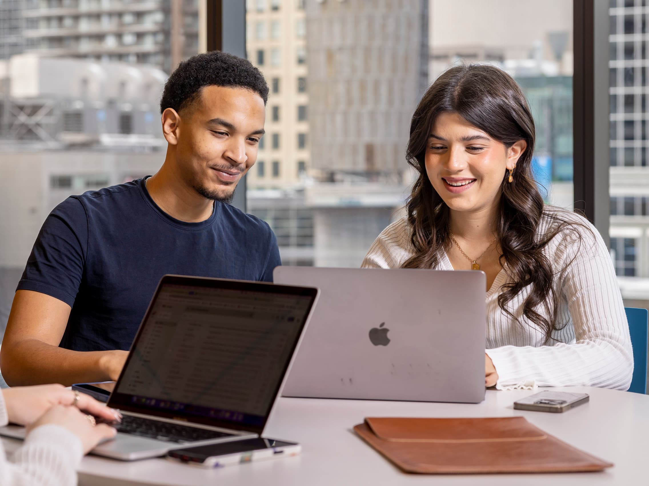 Two individuals sit at a table with laptops, smiling while working together. 