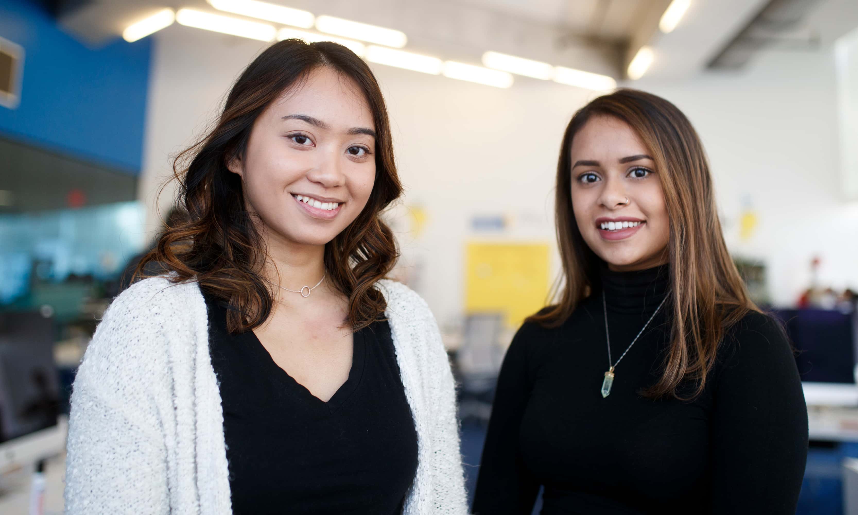 Two smiling women in dark tops, one with white cardigan, in a bright office with modern lighting.