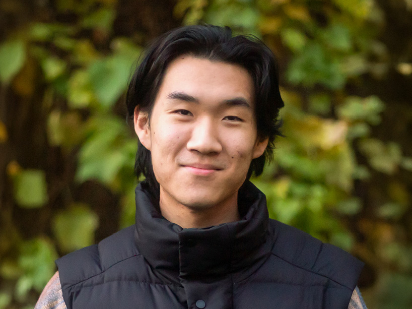 Person with dark hair smiles gently, wearing black vest against blurred green and yellow foliage outdoors.