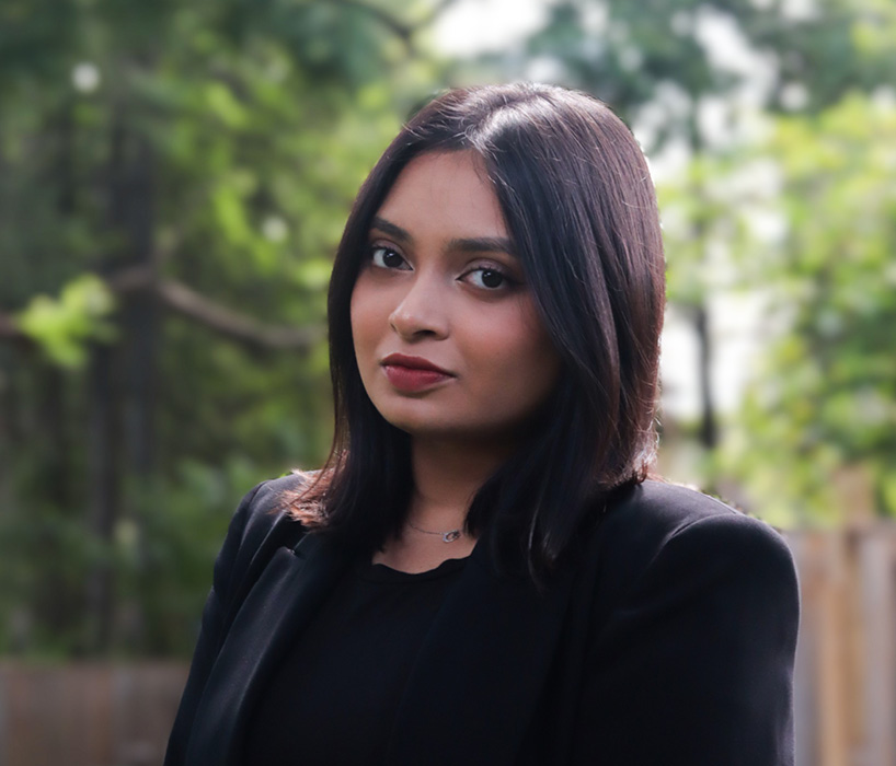 Woman with dark hair in black blazer stands outdoors, confidently facing camera with blurred greenery behind.