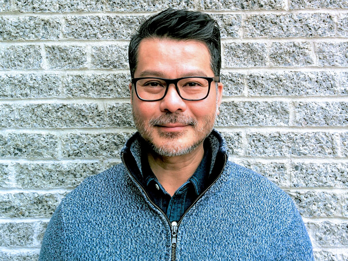 Man with glasses and beard smiles in blue sweater, standing by a textured brick wall.