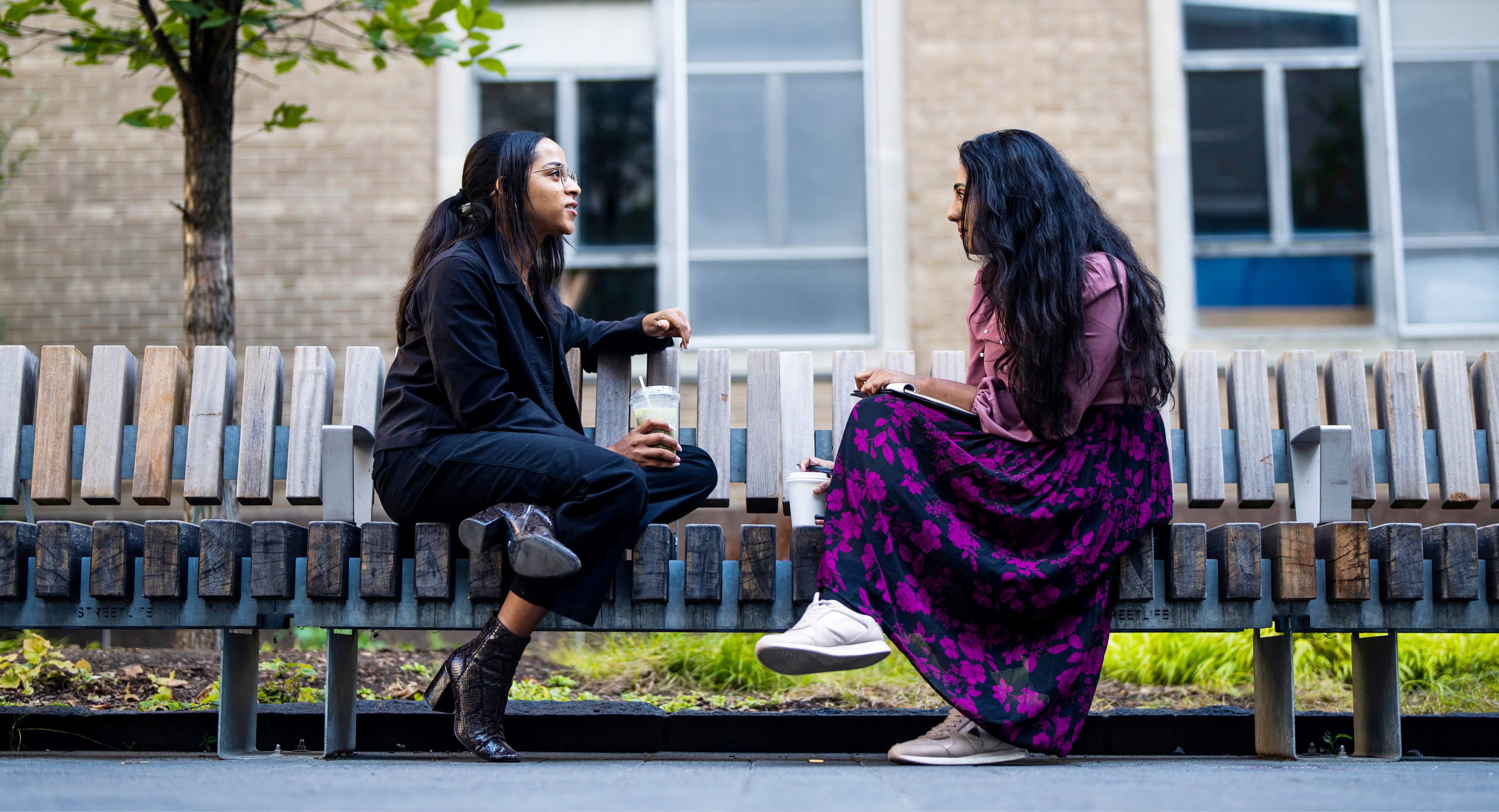 Two women chat on a bench outside. One in dark clothes, the other in a pink top and purple skirt.