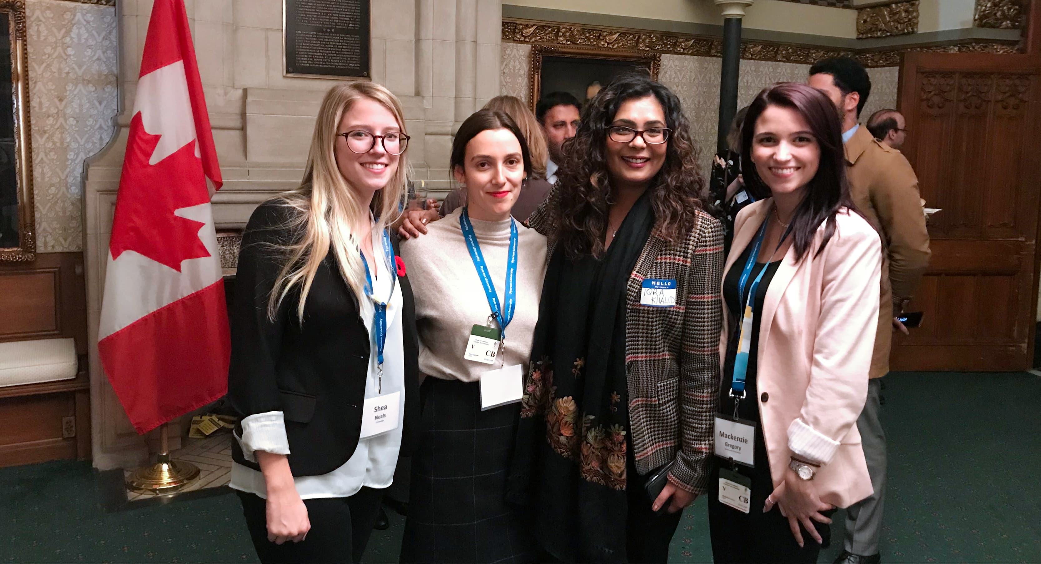 Four women in business attire smile in front of a large Canadian flag in the House of Commons..