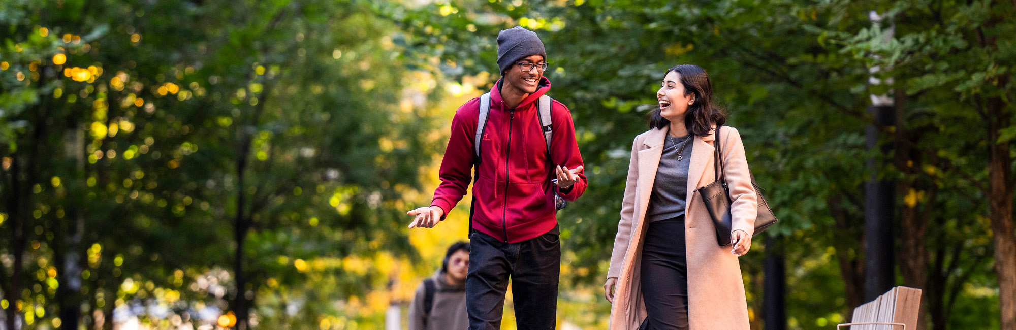 A man and woman chat joyfully, walking a leafy path under green trees.