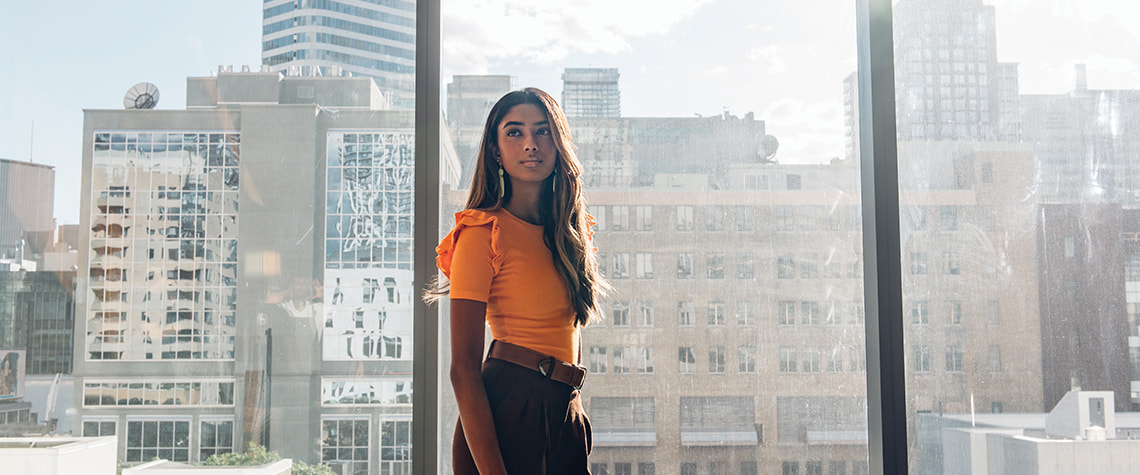 a female student standing in front of floor to ceiling windows with tall buildings in the background