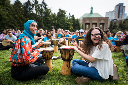 A candid photo of two, smiling female students drumming in the quad. In the background are more students drumming.