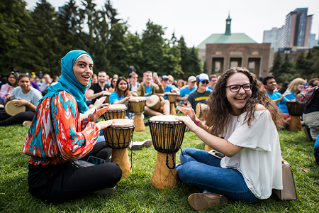 Students sitting on the grass participating in a drumming circle during an outdoor event.