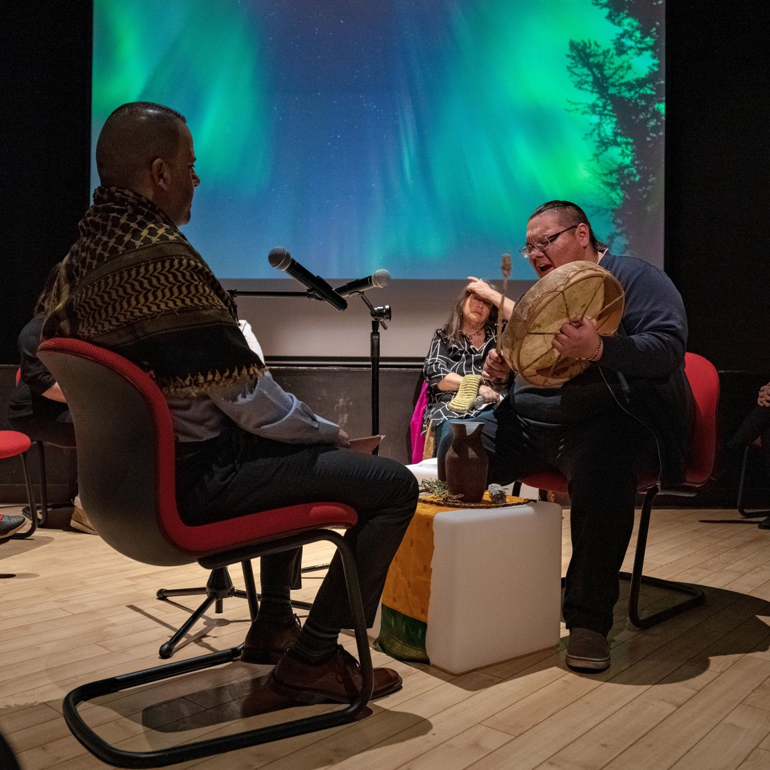 Two men sitting in front of each other, one playing a musical instrument, the other sitting in front of a microphone, during an art performance
