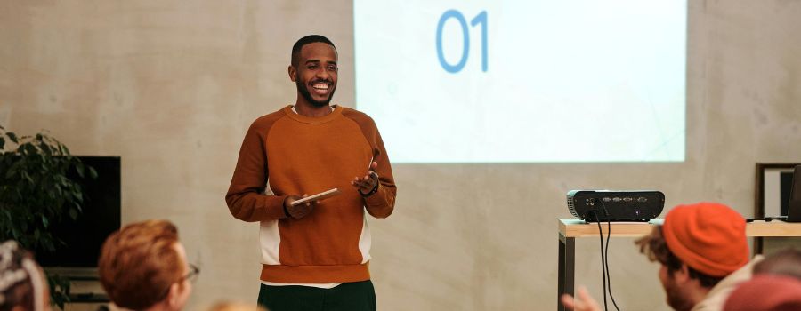 A Black man standing in front of a digital presentation, facing a group of people listening