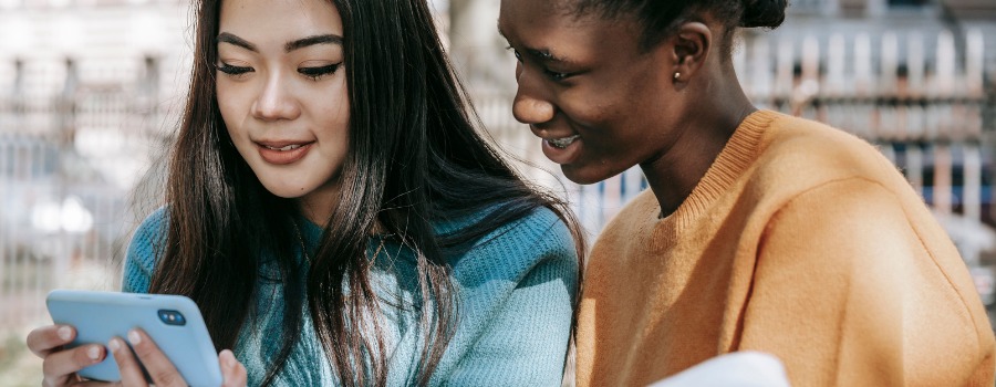 Two young girls, both racialized, looking at a phone and smiling