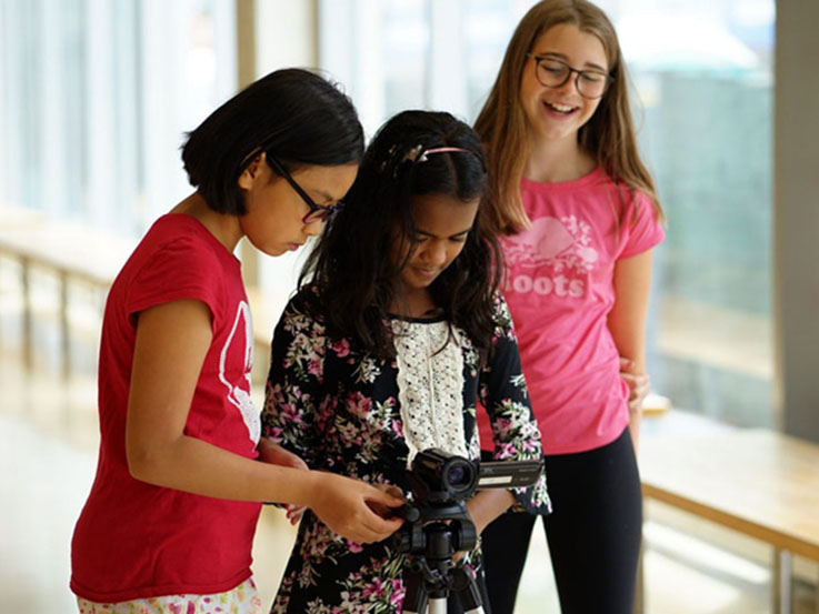 Three youth look down at the display screen of a video camera on a tripod