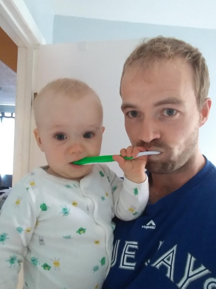 A man in a Blue Jays shirt holds an infant. The infant is grabbing the toothbrush from the man's mouth and is chewing on the end.
