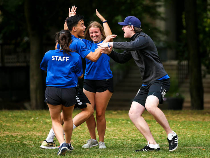 Three camp counsellors encircle a fourth holding exuberant facial expressions and body language.