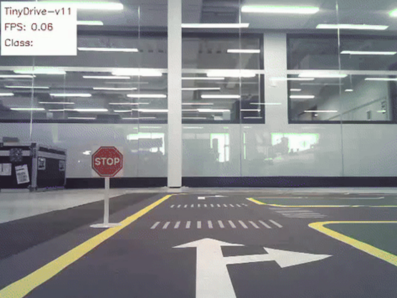 Indoor simulated driving track viewed from a low angle, featuring a miniature stop sign on the left, yellow lane markings, and directional arrows painted on dark grey pavement. A white overlay in the top-left displays software text and stats.
