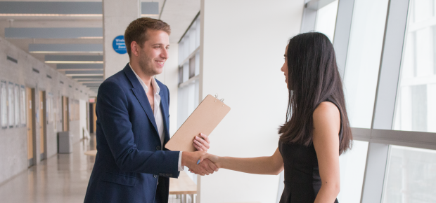 A man holding a folder shakes hands with a woman standing in a long, white corridor.