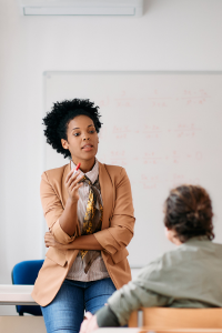 A lecturer sits on a desk while speaking to a class, with an observer show at the front of the room.