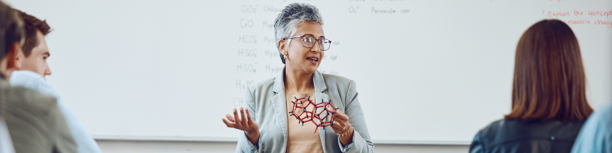 A lecturer shows students a plastic model of a molecule in front of a classroom whiteboard. 
