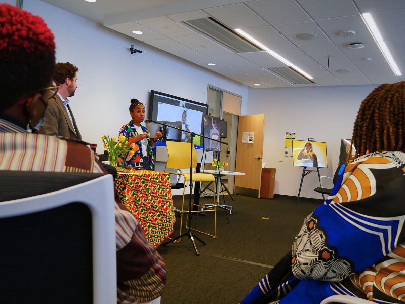 Two people sit in front of a classroom. One person speaks into a microphone as a group watches.