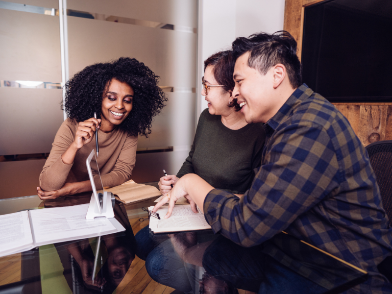 Three smiling people sit together at a table with books while one gestures towards a tablet. 