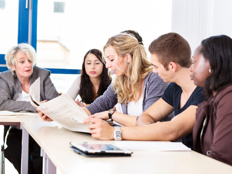 A group of six people sitting at a desk focused on one person who is reading from a paper.