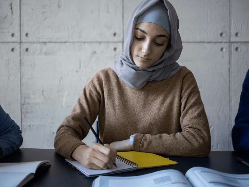 A person is facing the camera and sitting at a desk working with a pen and paper notepad.
