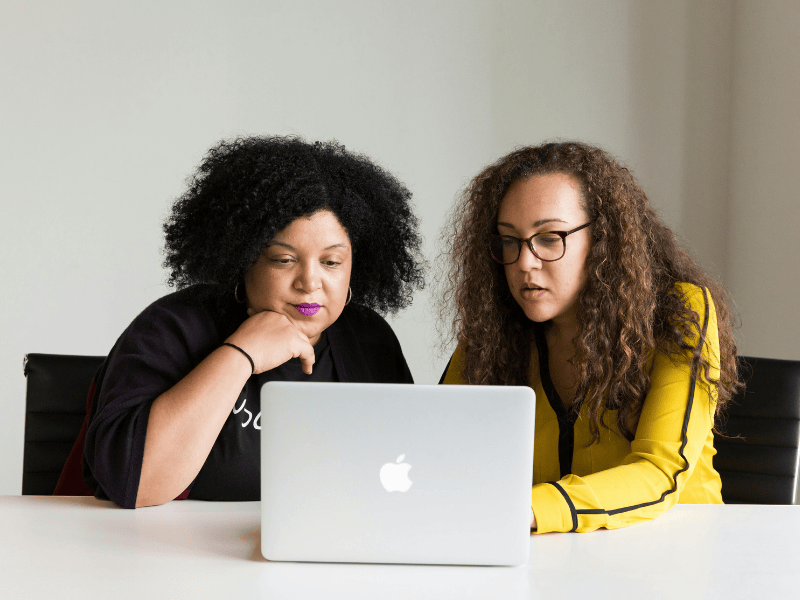 Two people sitting side by side facing the camera have a discussion while looking at a laptop screen.