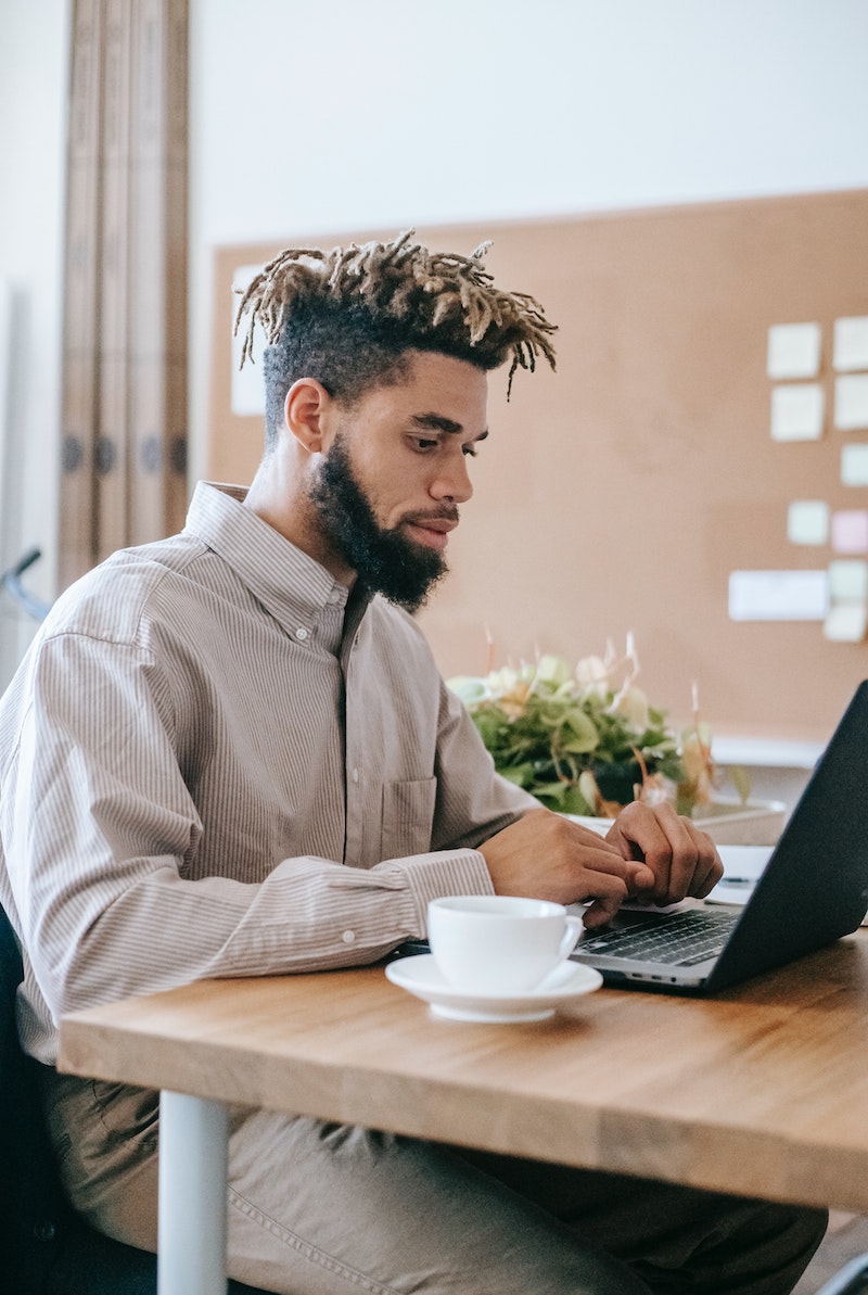 Man in beige shirt with short blonde dreadlocks works at a laptop with a mug