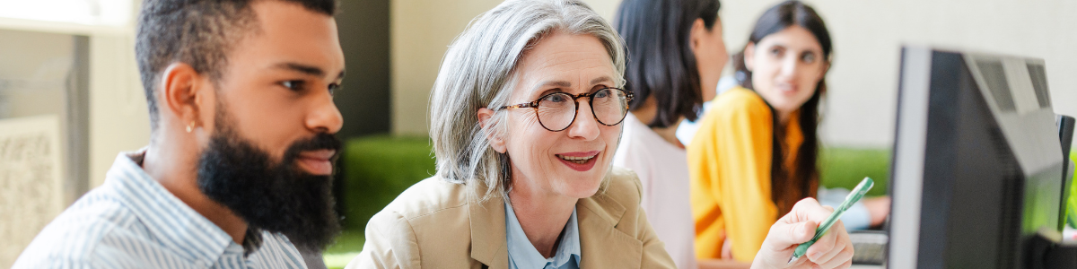 Two people sit in a classroom, working on a shared computer screen. One of them points with a pen while the other types on a keyboard. Another group works similarly in the background.