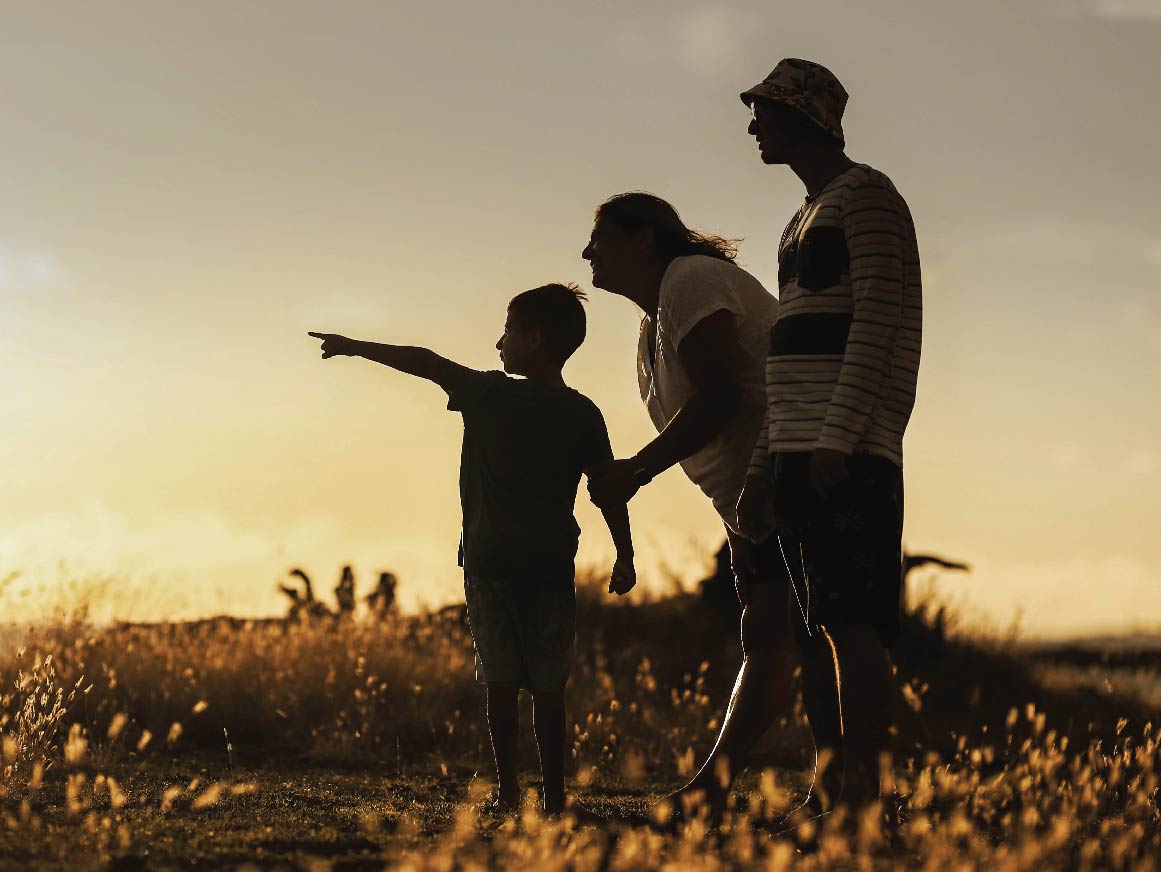 A small child, mother and father stand in nature looking at a sunset together