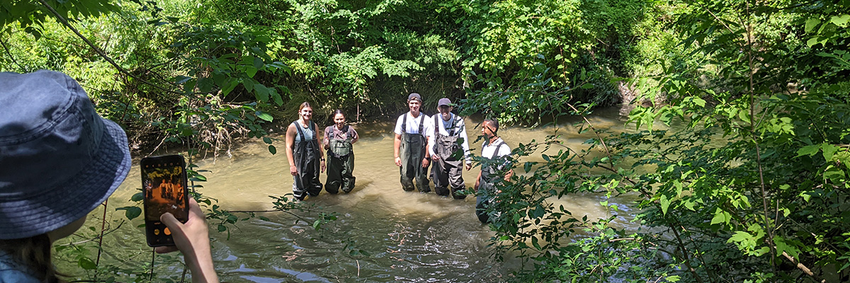 Student taking a photo of 5 students in overalls standing in a wetland