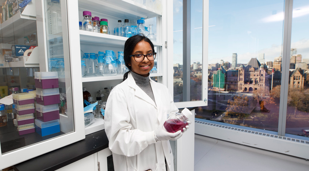 Chemistry and Biology graduate student holding a beaker with liquid in the MaRS lab, wtih a view of Queen's Park seen through window.