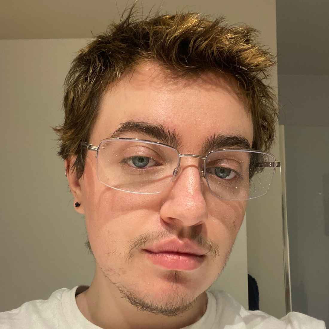 A young man with brown hair, blue eyes, wire-rimmed glasses, a small chin goatee, and a black stud earring, looking directly into a mirror with a neutral expression.