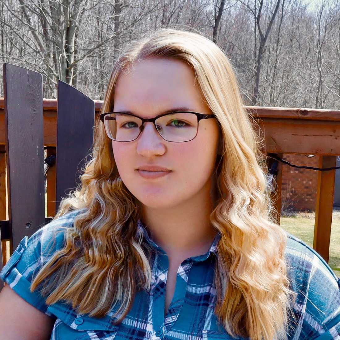 Young woman with wavy blonde hair and glasses, wearing a blue plaid shirt, sits pensively on a wooden deck with bare trees in the background.
