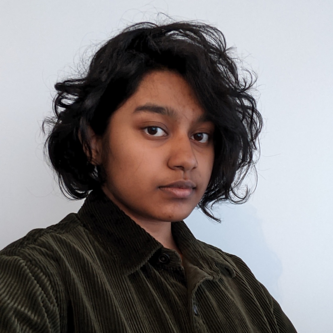  A person with wavy black hair and an olive corduroy shirt gazes calmly at the camera against a plain white background.