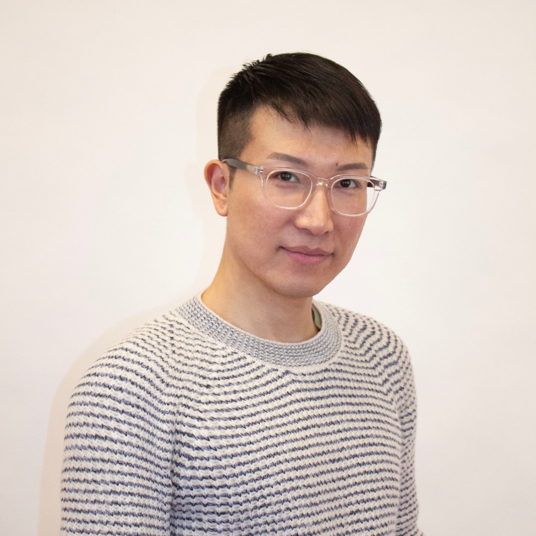 A man with short hair and glasses, wearing a striped sweater, stands against a plain background. He has a slight smile and looks directly at the camera.