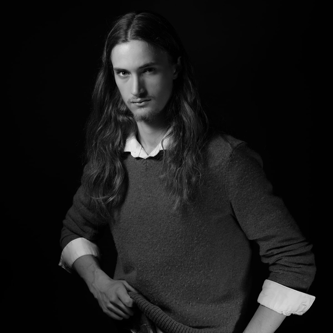 A young man with long hair and a goatee poses confidently in a black and white portrait. They wear a collared shirt under a sweater, set against a dark background.