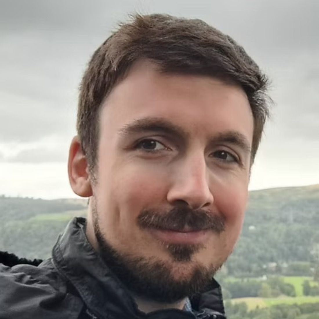 Portrait of a smiling man with dark hair and a beard, wearing a black jacket. He stands outdoors with a scenic, cloudy landscape in the background.