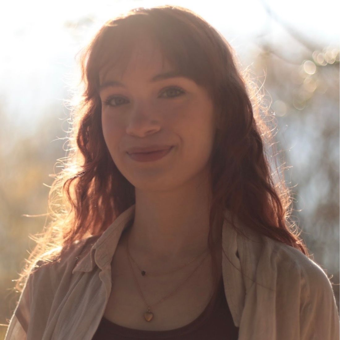 A smiling young woman with long, wavy reddish-brown hair and bangs, illuminated by warm, bright backlight outdoors.